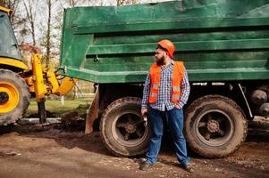 brutale baard werknemer man pak bouwvakker in oranje veiligheidshelm, tegen dump truck met hamer en verstelbare sleutel bij de hand. foto