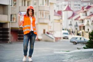 ingenieur bouwer vrouw in uniform vest en oranje beschermende helm houdt zakelijke tekenpapierrol vast tegen nieuwbouw. eigendom woonblok thema. foto