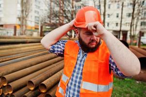 baard werknemer man pak bouwvakker in oranje veiligheidshelm in de buurt van stalen buizen. foto