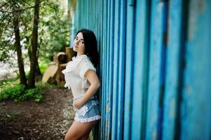 portret van sexy brunette meisje op vrouwen jeans shorts en witte blouse tegen blauwe houten huis met stompen. foto