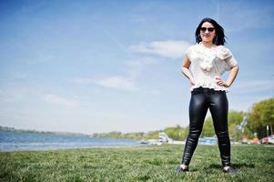 brunette meisje op vrouwen leren broek en witte blouse, zonnebril, gesteld op geen gras tegen het strand van het meer. foto