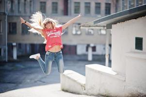 stijlvolle gelukkige blonde vrouw draagt jeans, zonnebrillen en t-shirt die bij zonnig weer op straat springen. mode stedelijk model portret. foto