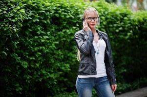 stijlvolle blonde vrouw draagt jeans, bril en leren jas met telefoon bij de hand, tegen struiken op straat. mode stedelijk model portret. foto