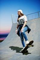 jonge stedelijke tienermeisje met skateboard, draag een bril, pet en gescheurde spijkerbroek in het skatepark op de avond. foto