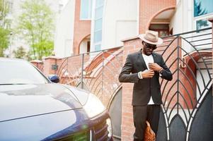 stijlvolle zwarte man met bril met hoed, draag pak met handtas tegen luxe auto. rijke Afro-Amerikaanse zakenman. foto