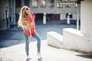 stijlvolle gelukkige blonde vrouw draagt jeans, zonnebrillen en t-shirt op straat bij zonnig weer. mode stedelijk model portret. foto