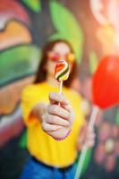 mooie tiener die lolly toont, bril draagt, hartballon bij de hand, geel t-shirt in de buurt van graffitimuur. foto