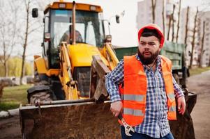 brute baard werknemer man pak bouwvakker in oranje veiligheidshelm, tegen traktor met verstelbare sleutel bij de hand. foto