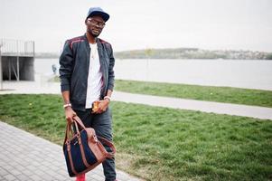 portret van stijlvolle Afro-Amerikaanse man op sportkleding, pet en bril wandelen met handtas. zwarte mannen model street fashion. foto