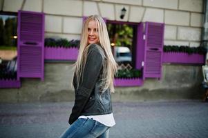 stijlvolle blonde vrouw draagt spijkerbroek en jas op straat tegen paars raam. mode stedelijk model portret. foto