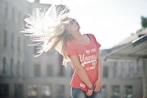 stijlvolle gelukkige blonde vrouw draagt jeans, zonnebrillen en t-shirt op straat bij zonnig weer. mode stedelijk model portret. foto