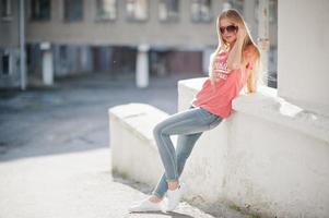 stijlvolle gelukkige blonde vrouw draagt jeans, zonnebrillen en t-shirt op straat bij zonnig weer. mode stedelijk model portret. foto