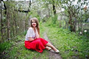 portret van een mooi meisje met rode lippen in de lentebloesemtuin op groen gras, een rode jurk en een witte blouse dragen. foto