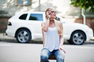 stijlvolle blonde vrouw draagt jeans, bril, choker en wit overhemd in de buurt van stoel tegen luxe auto. mode stedelijk model portret. foto