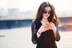 portret brunette meisje met rode lippen en oranje mobiele telefoon bij de hand, gekleed in een zwarte jurk, zonnebril op het dak gesteld. straatmodel. foto