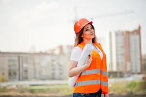 ingenieur bouwer vrouw in uniform vest en oranje beschermende helm houdt zakelijk papier tegen nieuwe gebouwen met kraan. foto