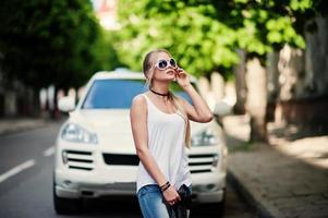 stijlvolle blonde vrouw draagt jeans, zonnebrillen, choker en wit overhemd tegen luxe auto. mode stedelijk model portret. foto