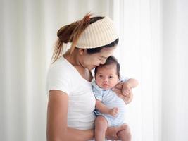 gelukkige aziatische moeder die een mooie pasgeboren baby vasthoudt in de kinderkamer, moeder die haar pasgeboren baby knuffelt en bij het raam staat foto