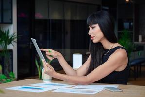 Aantrekkelijke blanke zakenvrouw die een tabletcomputer gebruikt om gegevens te zoeken terwijl ze op de tafel zit met een informatiedocument en werkt op een modern kantoor foto