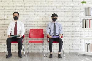 twee aziatische mannen zitten in rode plastic stoelen met mat uit elkaar geplaatst om hun mond te bedekken om covid-19 te voorkomen terwijl ze wachten op een sollicitatiegesprek. foto