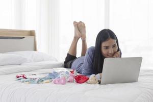 een aziatische moeder is op een laptop op zoek naar babykleding voor haar kind, online winkelend in bed met een stralende glimlach. foto