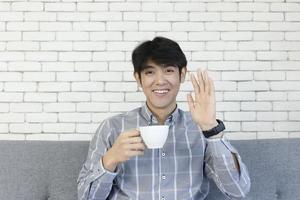 een jonge aziatische man, met een koffiekopje en glimlachend zwaaiend met zijn hand. foto
