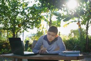 jongen die online studeert, houdt een pen vast om op een tablet te schrijven en kijkt naar het laptopscherm met de ochtendzon achter hem in een landhuis. concept online onderwijs platteland foto