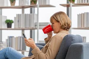 ontspannende vrouw met kopjes koffie in de woonkamer, volwassen vrouw die mobiele smartphone gebruikt tijdens een koffiepauze foto
