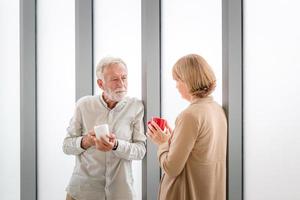 Senior paar in nieuw huis tijdens koffiepauze, ouder echtpaar praten terwijl ze bij het raam staan met kopjes koffie foto