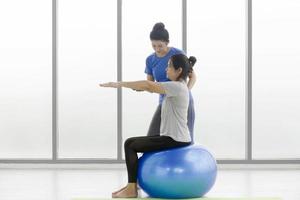 een vrouwelijke trainer leert een Aziatische vrouwelijke klant van middelbare leeftijd om yoga te doen, zittend op een rubberen bal in een gymnasium. foto