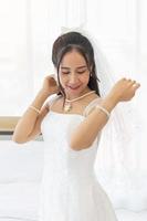 een aziatische bruid gekleed in een witte trouwjurk staat in een schattige en heldere glimlach naast het bed in de kamer. foto