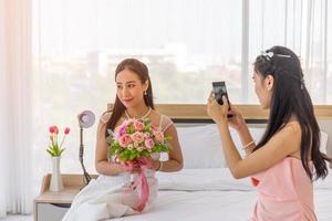 de vriendin van de bruid gebruikt een smartphone om een foto te maken van een Aziatische bruid in een trouwjurk in haar hand, met een groot, mooi boeket bloemen op het bed.