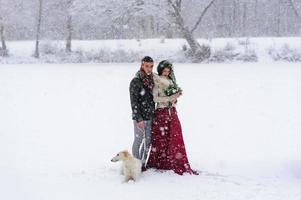 mooie bruid en bruidegom met een witte hond staan op de achtergrond van een besneeuwd bos. foto