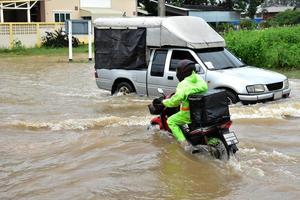 bezorging aziatische man met groen uniform, rijdend en etend bezorgend aan klant met voedselkoffer achter in overstromingsstraat, expresbezorging van voedsel en online winkelen concept in de overstromingssituatie. foto