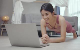 jonge sterke vrouw traint thuis, staat in plankpositie, traint met online trainer met behulp van laptop. foto