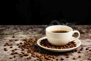 close-up van warme koffie in een witte kop wordt op een betonnen vloertafel geplaatst, veel gebrande koffiebonen zitten in een koffiekopschotel en rond, rook en aroma waaien uit de kop. zwarte, onscherpe achtergrond