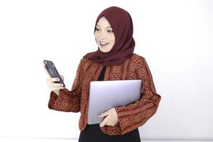 jonge aziatische islamvrouw staat en lacht als ze aan de telefoon kijkt terwijl ze een laptop vasthoudt foto