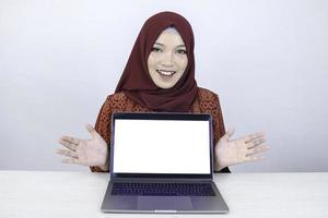 jonge aziatische islamvrouw lacht wijzend en toont op een wit leeg scherm op de laptop naar de camera. foto