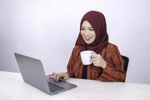 mooie jonge aziatische islamvrouw werkt graag op een laptop terwijl ze in de witte kamer zit en koffie drinkt. foto