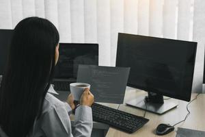 Aziatische vrouw dronk koffie in de vroege ochtend terwijl ze het programma en de code op het computerscherm overwoog. foto