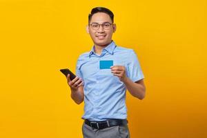 portret van lachende jonge Aziatische man aziatisch in glazen met mobiele telefoon en creditcard geïsoleerd op gele achtergrond. zakenman en ondernemer concept foto