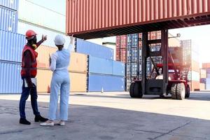 achter twee arbeiders dragen een veiligheidsvest en helm die discussiëren op de logistieke vrachtcontainerwerf. Afro-Amerikaanse ingenieur man praten met mooie jonge vrouw baas met blond haar op de werkplek. foto