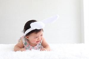 portret van een zes maanden kruipende baby op pluizig wit tapijt, schattig lief klein meisje met konijnenoren hoofdband huilend op bed in slaapkamer, kindertijd en Pasen decoratie concept foto