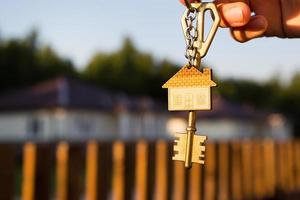 sleutelhanger met sleutelhuis in de hand. achtergrond van hek en huisje. verhuizen naar een nieuw huis, hypotheek, onroerend goed kopen, woningen huren en boeken, dromen van wonen in het dorp. kopieer ruimte foto