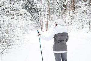 skiër een vrouw in een membraanjas met skistokken in zijn handen met zijn rug tegen de achtergrond van een besneeuwd bos. langlaufen in winterbos, buitensporten, gezonde levensstijl. foto