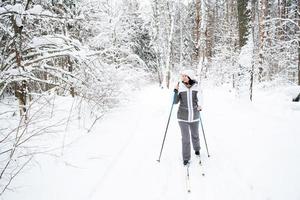 skiër een vrouw in een membraanjas met skistokken in zijn handen met zijn rug tegen de achtergrond van een besneeuwd bos. langlaufen in het winterbos, buitensporten, gezonde levensstijl. foto
