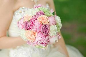 huwelijksboeket met roze en perzikbloemen in de handen van de bruid. groene anjer, paarse roos, witte pioen foto