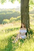 portret van een jong mooi meisje in een zomerjurk. zomer fotosessie in het park bij zonsondergang. een meisje zit onder een boom in de schaduw. foto