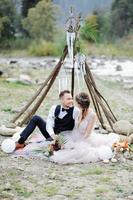 aantrekkelijk paar pasgetrouwden, gelukkig en vreugdevol moment. man en vrouw in feestelijke kleding zitten op de stenen in de buurt van de huwelijksdecoratie in boho-stijl. ceremonie buiten. foto