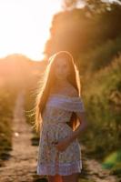 portret van een jong mooi meisje in een zomerjurk. zomer fotosessie in het park bij zonsondergang. een meisje zit onder een boom in de schaduw. foto